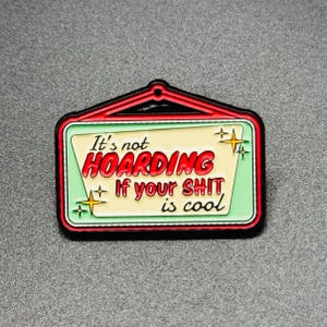 May include: A rectangular enamel pin with a black border and a red outline. The pin features the text "It's not HOARDING If your SHIT is cool" in red and white, with star accents on a light green and cream background.