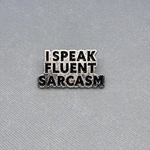 Könnte beinhalten: Silberfarbener Pin mit schwarzem Text, der "Ich spreche fließend Sarkasmus" lautet.