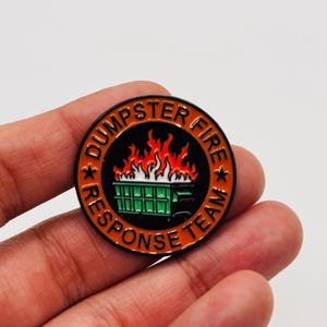 May include: A round enamel pin with the words "DUMPSTER FIRE RESPONSE TEAM" around the edge. The center features a green dumpster engulfed in flames. The pin has a black border with orange and white accents.