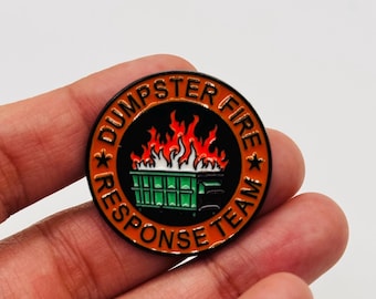 May include: A round enamel pin with the words "DUMPSTER FIRE RESPONSE TEAM" around the edge. The center features a green dumpster engulfed in flames. The pin has a black border with orange and white accents.