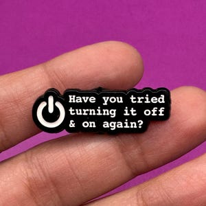 Puede incluir: Un charm negro y blanco con el texto "Have you tried turning it off & on again?" y un símbolo de botón de encendido. El charm es rectangular con bordes redondeados, sobre un fondo morado.