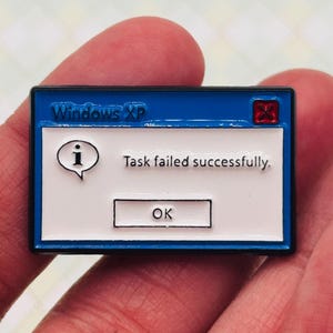 Könnte beinhalten: Ein blau-weißer Emaille-Pin mit einer Windows XP-Fehlermeldung. Der Pin zeigt oben "Windows XP" und "Task failed successfully." mit einer "OK"-Schaltfläche darunter. Ein rotes "X" befindet sich in der oberen rechten Ecke.