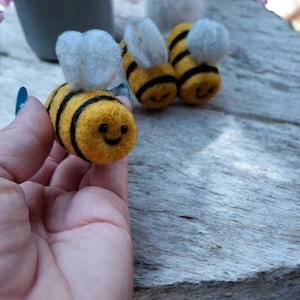 May include: Three small needle-felted bees with yellow and black striped bodies, white fuzzy wings, and stitched black eyes and smiles. Each bee has a rounded, textured wool appearance, with one bee held in the foreground and two others behind.
