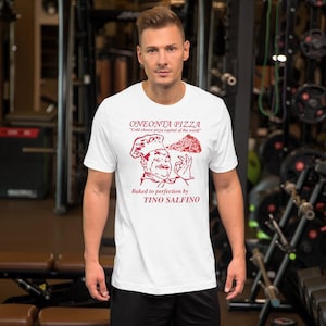 May include: White t-shirt with a red graphic featuring the text "ONEONTA PIZZA" and "Cold cheese pizza capital of the world." The graphic includes an illustration of a chef holding a pizza slice, with the text "Baked to perfection by TINO SALFINO."