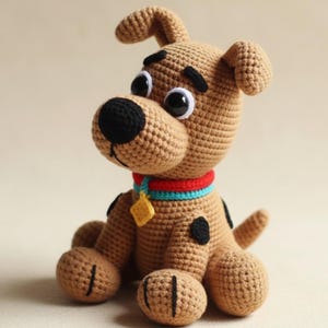 Puede incluir: Un perro de ganchillo color canela con manchas negras, una nariz negra y ojos grandes negros y blancos. El perro lleva un collar rojo y azul con una etiqueta dorada. El peluche hecho a mano está sentado.