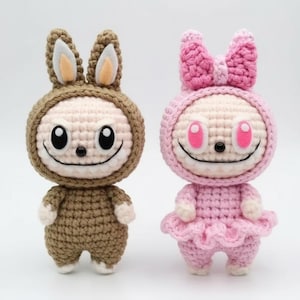 May include: Two handmade crochet dolls. One is brown with bunny ears, the other is pink with a bow and a skirt. Both dolls have smiling faces with large black eyes and white teeth. The dolls are standing upright.