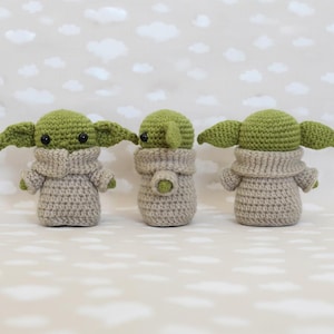 May include: Three handmade crochet figures resembling a popular character. Each figure has large green ears, black eyes, and is wearing a tan-colored outfit. The figures are displayed against a backdrop of a light gray background with white cloud shapes.