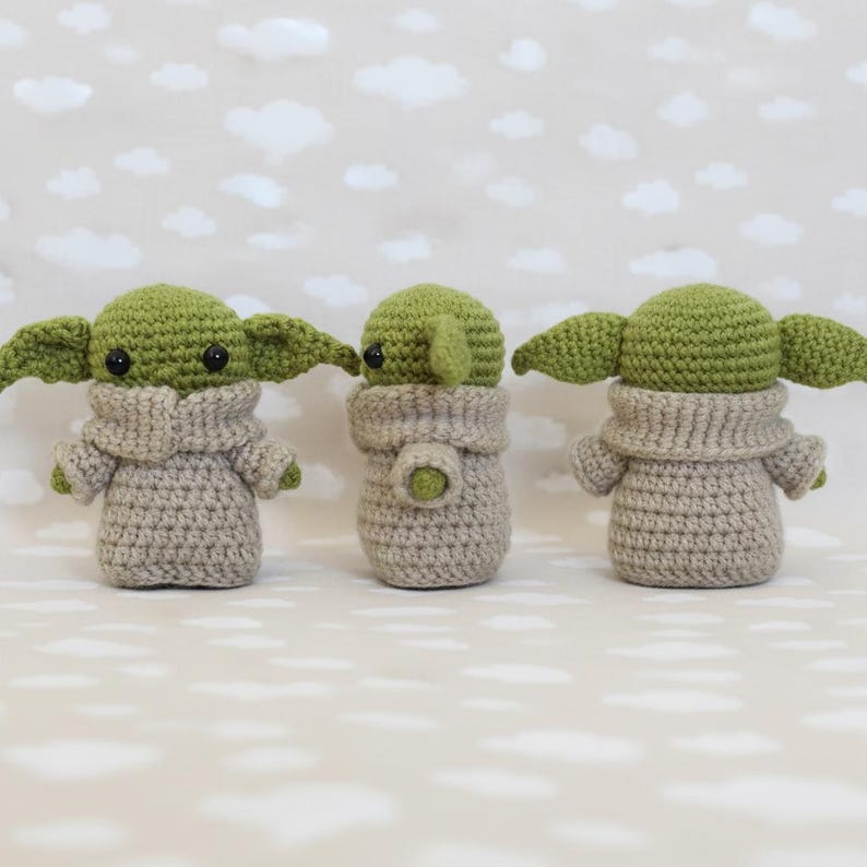 May include: Three handmade crochet figures resembling a popular character. Each figure has large green ears, black eyes, and is wearing a tan-colored outfit. The figures are displayed against a backdrop of a light gray background with white cloud shapes.