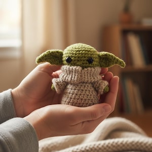 May include: A handmade crochet doll of a character with large ears and black eyes. The doll is green and tan, with a tan robe and a green head. The doll is held in two hands.