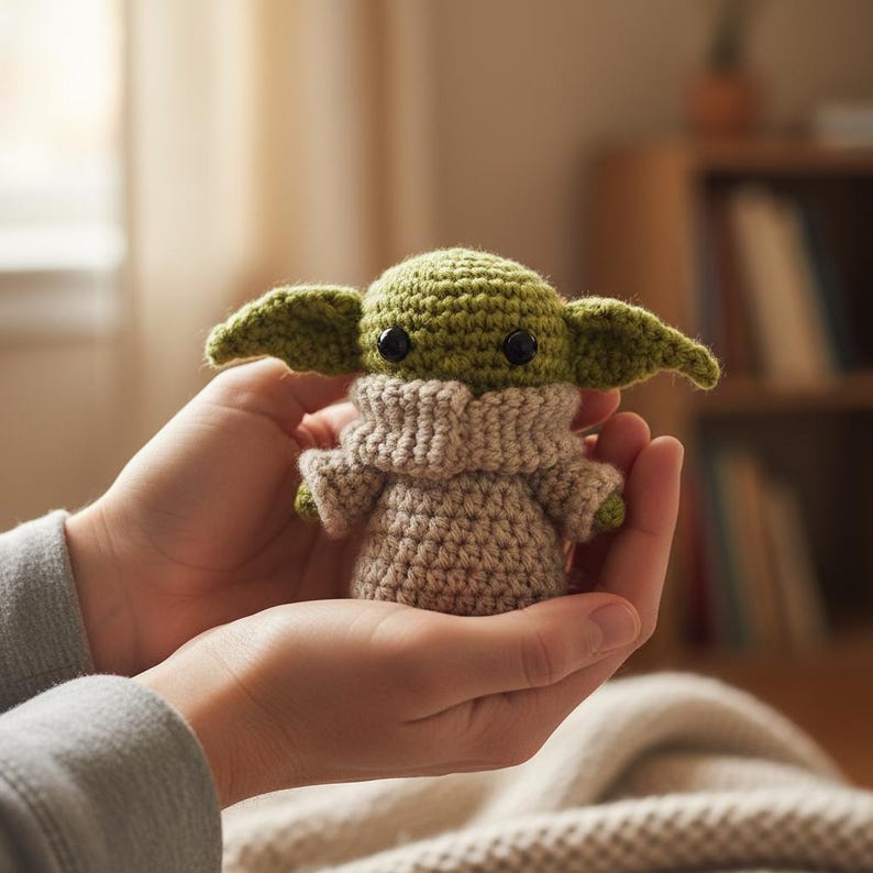 May include: A handmade crochet doll of a character with large ears and black eyes. The doll is green and tan, with a tan robe and a green head. The doll is held in two hands.