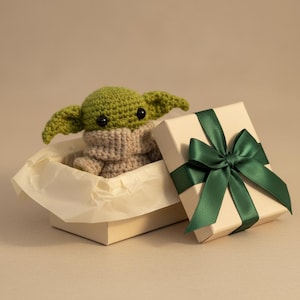 May include: A crocheted green and tan character sits inside an open gift box with a green satin ribbon. The character has large ears and black eyes. The gift box is off-white and lined with tissue paper.