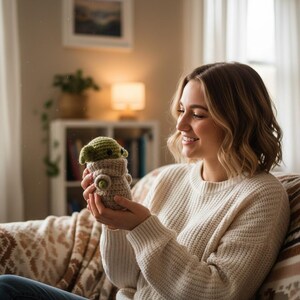 May include: A handmade crochet doll of a character with large ears and a tan and green color scheme. The doll is held in a person's hands. The doll is wearing a tan outfit with green accents. The background is a cozy living room setting.