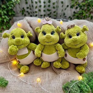 May include: Three handmade green crochet dolls, resembling a popular animated character, are displayed. Each doll has a smiling face, black eyes, and a cream-colored diaper. One doll has a pink bow, and another has a brown hair accessory.