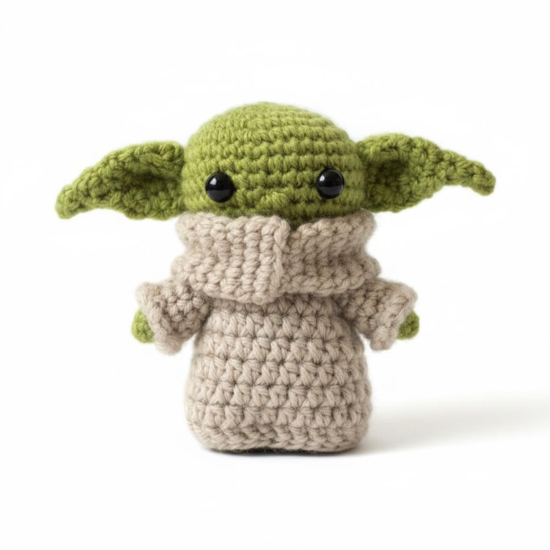 May include: A handmade crochet doll featuring a green head and large ears, black eyes, and a tan robe. The doll is made of yarn and has a rounded body. The overall design is inspired by a popular character from a science fiction franchise.