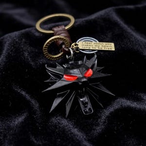 May include: Black metal keychain featuring a wolf head with red eyes. The keychain includes a small tag with the inscription "I feel about you makes my heart long to be free." The keychain has a brown leather strap and a gold-colored ring.
