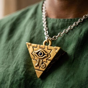 May include: A gold-coloured pendant necklace with a silver chain. The pendant is triangular with an eye design at its centre. The eye is blue with gold detailing. The necklace is worn over a green shirt.