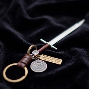 May include: A silver sword-shaped keychain with a brown handle and a gold-colored keyring. A small gold tag with the text "What is the poin makes my heart love in so free" hangs from the keychain.