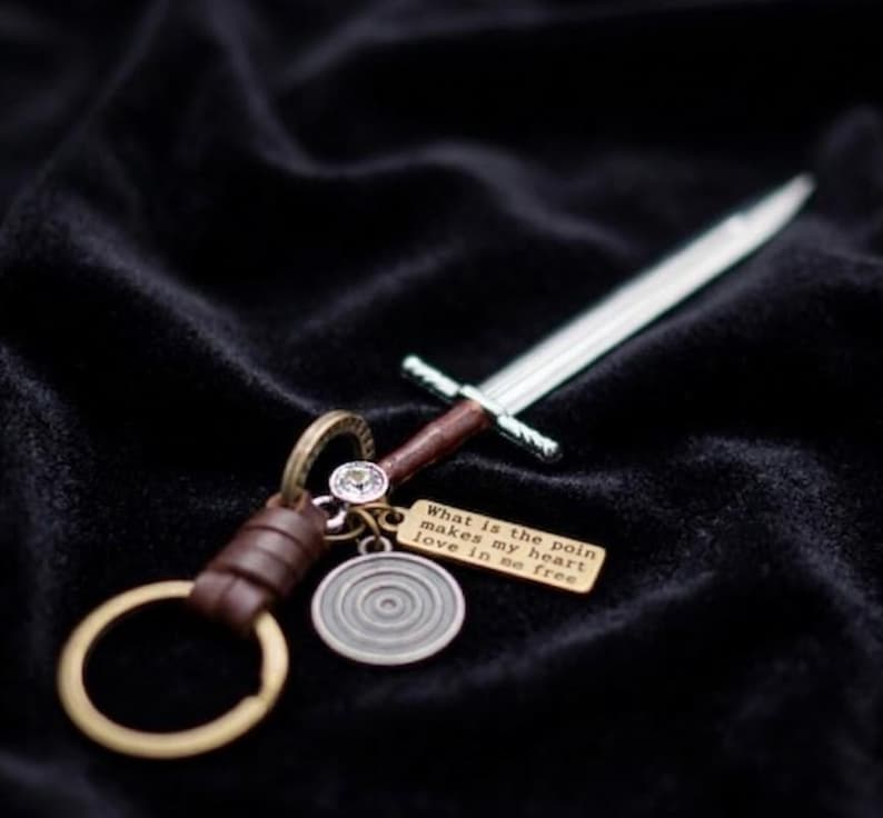 May include: A silver sword-shaped keychain with a brown handle and a gold-colored keyring. A small gold tag with the text "What is the poin makes my heart love in so free" hangs from the keychain.