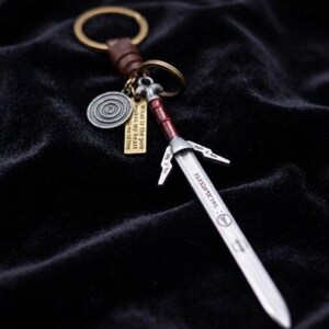 May include: A silver sword keychain with a red handle and a brown leather strap. The keychain includes a circular charm and a small tag with text. The sword has engraved text on the blade.