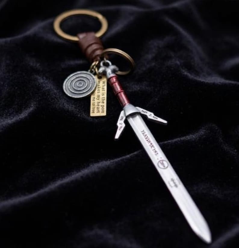 May include: A silver sword keychain with a red handle and a brown leather strap. The keychain includes a circular charm and a small tag with text. The sword has engraved text on the blade.