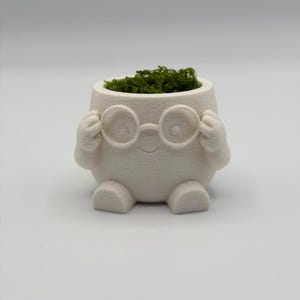 May include: A white, round, 3D-printed planter shaped like a character with small feet and arms holding a pair of round glasses. The planter features a smooth, matte finish and contains a small amount of bright green moss inside the top opening.