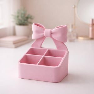 Pode incluir: Um organizador rosa claro com quatro compartimentos, encimado por um laço decorativo. O laço é da mesma cor que o organizador. O organizador está sobre uma superfície branca.