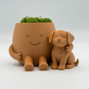 May include: A light brown 3D-printed planter shaped like a seated figure with a simple smiling face, filled with green moss. Beside the planter sits a matching light brown 3D-printed puppy figurine with a collar, being hugged by the planter figure.