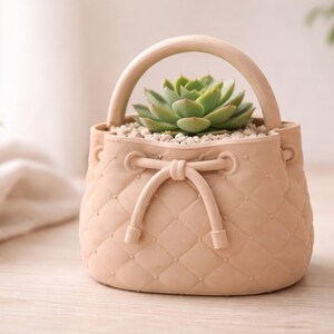 May include: A peach-colored, purse-shaped planter with a handle, filled with small white stones and a green succulent. The planter has a quilted pattern and a decorative bow. A charming decorative item.