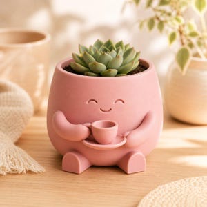 May include: A pink ceramic planter shaped like a smiling character, holding a small teacup and saucer. The planter is filled with soil and a green succulent plant. The design is whimsical and decorative.