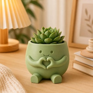 May include: A green ceramic planter shaped like a character with a smiling face and arms forming a heart shape. The planter holds a green succulent plant. The planter is on a wooden surface.