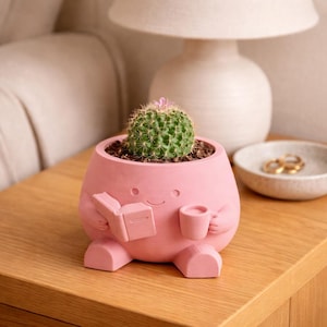 May include: A pink, cartoon-like planter with a smiling face, holding a book and a cup. The planter contains a small cactus with green needles and a pink flower. The planter is sitting on a wooden surface.