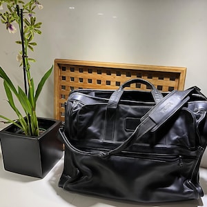 Pode incluir: Pasta de couro preta com alça de ombro, com a marca "TUMI". A bolsa tem vários compartimentos e uma alça superior. À esquerda, há uma planta em vaso com folhas verdes e flores brancas e roxas.