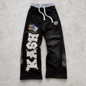 May include: Black sweatpants featuring a white drawstring waist, a large white distressed-style patch reading KASH with star accents, flame graphics on the sides, and a small embroidered circular logo on the upper left thigh.
