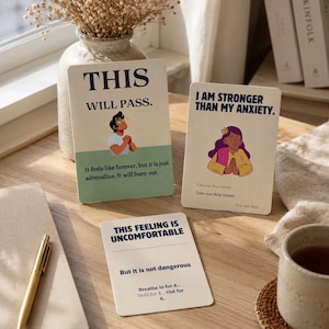 May include: Three rectangular affirmation cards with cream backgrounds and dark blue text. One card reads THIS WILL PASS, another I AM STRONGER THAN MY ANXIETY, and the third THIS FEELING IS UNCOMFORTABLE, each featuring illustrated figures and breathing instructions.