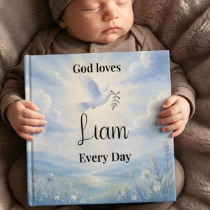 May include: A square hardcover book with a light blue sky background featuring a white dove carrying an olive branch. The text reads God loves Liam Every Day in black lettering. Below the text is a landscape illustration of rolling hills and flowers.