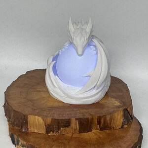 May include: A white dragon sculpture cradling a glowing blue orb. The dragon's head is detailed with horns and a textured face. The sculpture sits on a rustic, wooden base, creating a fantasy-themed decorative piece.