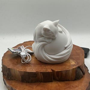 May include: A white cat sculpture curled up in a ball, resting on a wooden base. The cat's form is smooth and flowing, with a pointed ear and a curled tail. A charging device with a white cord is next to the sculpture.