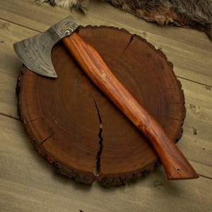 May include: A handcrafted axe with a patterned steel blade and a polished wooden handle. The axe is resting on a dark brown wooden cross-section, showcasing the wood grain. The handle features brass accents.