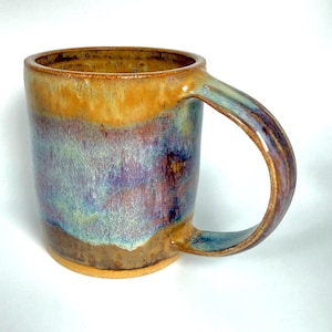 May include: A handmade ceramic mug with a unique glaze. The mug features a blend of colors, including shades of orange, purple, blue, and brown. The handle is curved and integrated into the mug's design. The mug is suitable for hot or cold beverages.