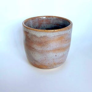 May include: A handmade ceramic cup with a cylindrical shape. The cup features a blend of brown, tan, and gray hues, creating a marbled effect. The rim is a darker brown. This stoneware cup is suitable for beverages.