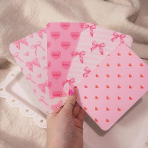 May include: A hand displays a selection of pink cards with Valentine's Day designs. The cards showcase patterns of hearts, bows, and the text "xoxo". The cards are presented on a white tray with a scalloped edge.