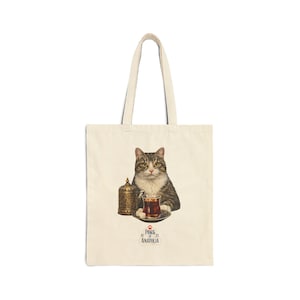 May include: Cream-colored tote bag featuring a cat with a tea cup and a decorative container. The cat is brown and gray with yellow eyes. The bag has a handle and the text "PAWS 1923 ANATOLIA".