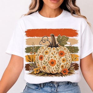 Peut inclure: Un t-shirt blanc à manches courtes avec une impression graphique représentant une citrouille décorée de fleurs orange et jaunes. La citrouille est placée sur un fond de bandes horizontales usées dans des tons orange, beige, vert et marron.