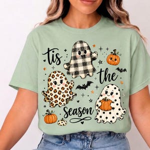 Peut inclure: Un t-shirt vert clair avec une impression graphique de trois fantômes de dessin animé aux motifs variés : carreaux vichy noir et blanc, imprimé léopard et pois noirs. Le design inclut des citrouilles orange, des chauves-souris et le texte 'tis the season'.