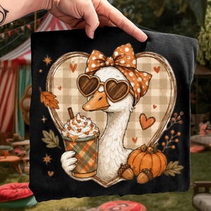 May include: A black t-shirt featuring a graphic of a white goose wearing heart-shaped sunglasses and an orange polka-dot bow. The goose holds a plaid cup with whipped cream, surrounded by a heart-shaped plaid frame, a pumpkin, and acorns.