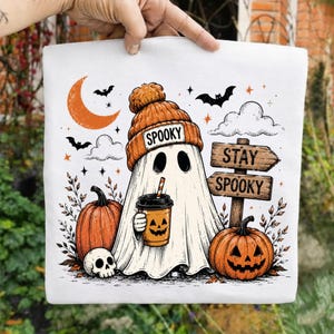 Peut inclure: Un sac fourre-tout blanc avec une illustration de fantôme portant un bonnet orange marqué SPOOKY, tenant un gobelet à café avec un visage de citrouille. Le motif inclut deux citrouilles, un crâne, un croissant de lune, des chauves-souris et un panneau.
