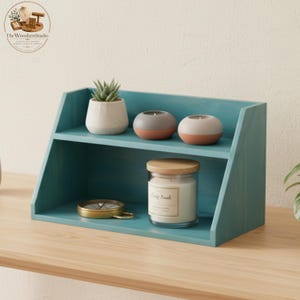 May include: A teal wooden shelf unit with two levels. The top shelf holds a small succulent in a white pot and two round candles. The bottom shelf displays a glass jar candle and a gold compass.