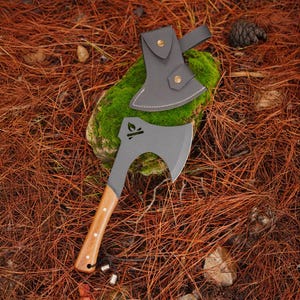 May include: A matte grey steel hatchet with a light brown wooden handle featuring three silver rivets and a small leaf icon cutout on the blade. The hatchet includes a matching grey protective sheath with two brass snap buttons for the blade edge.