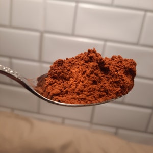 May include: A close-up of a spoon filled with a heaping amount of reddish-brown powder. The powder is finely ground and has a uniform texture.