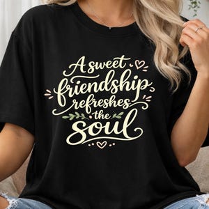 May include: Black t-shirt featuring the phrase "A sweet friendship refreshes the soul" in a cursive script. The design incorporates hearts and floral elements in light pink and green. The top has short sleeves.
